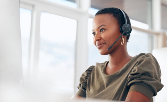 A young, black woman with a small smile is wearing a phone headset, olive-colored blouse and gold earrings.